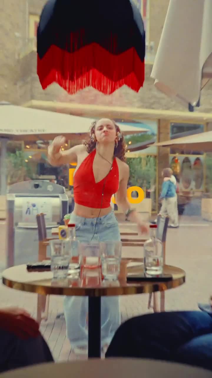 A girl dancing to Brazilian funk outside a restaurant while two people remain seated inside
